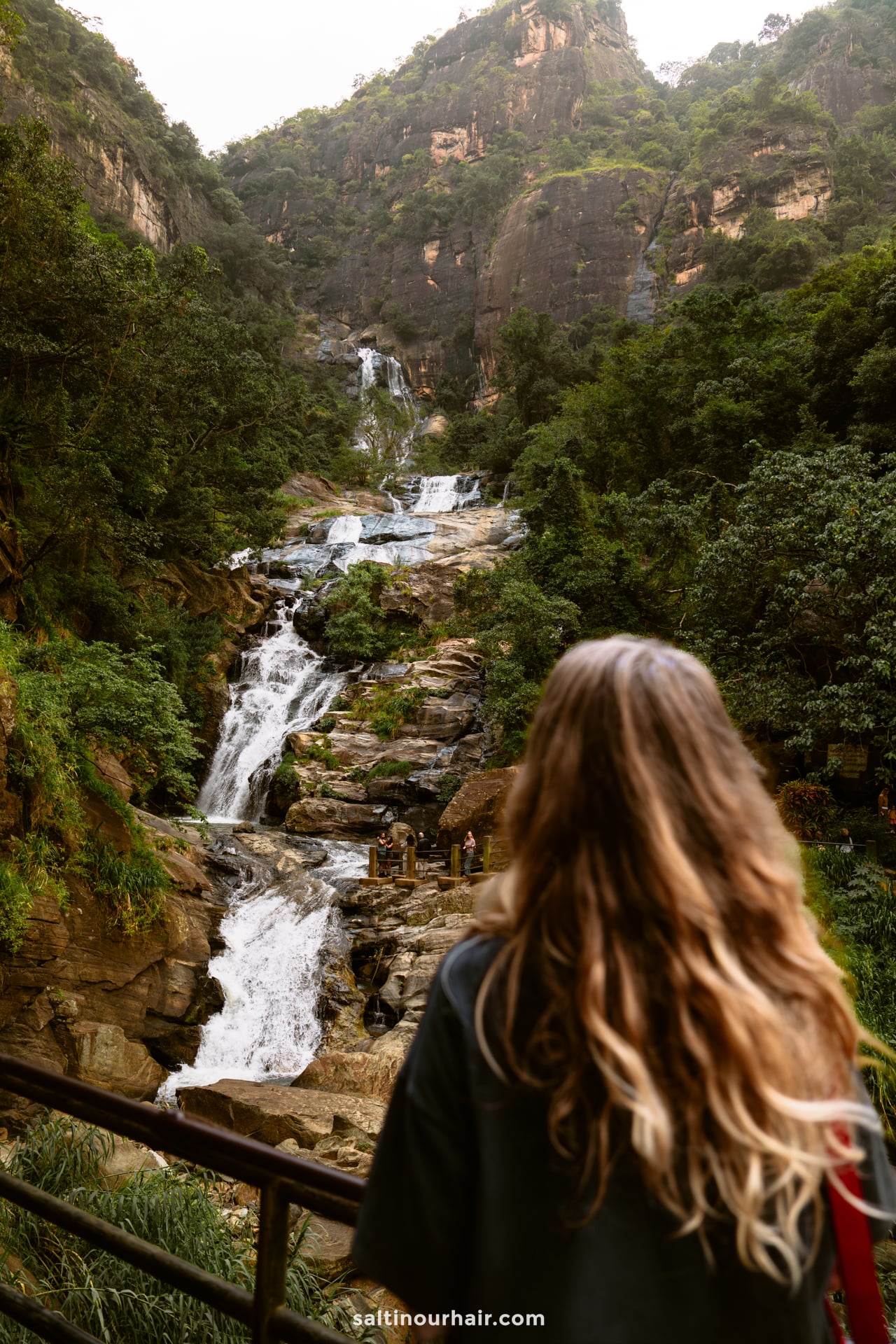 A person with long hair stands on a railing, gazing at a multi-tiered waterfall cascading down a rocky, forested mountain in Ella, Sri Lanka.