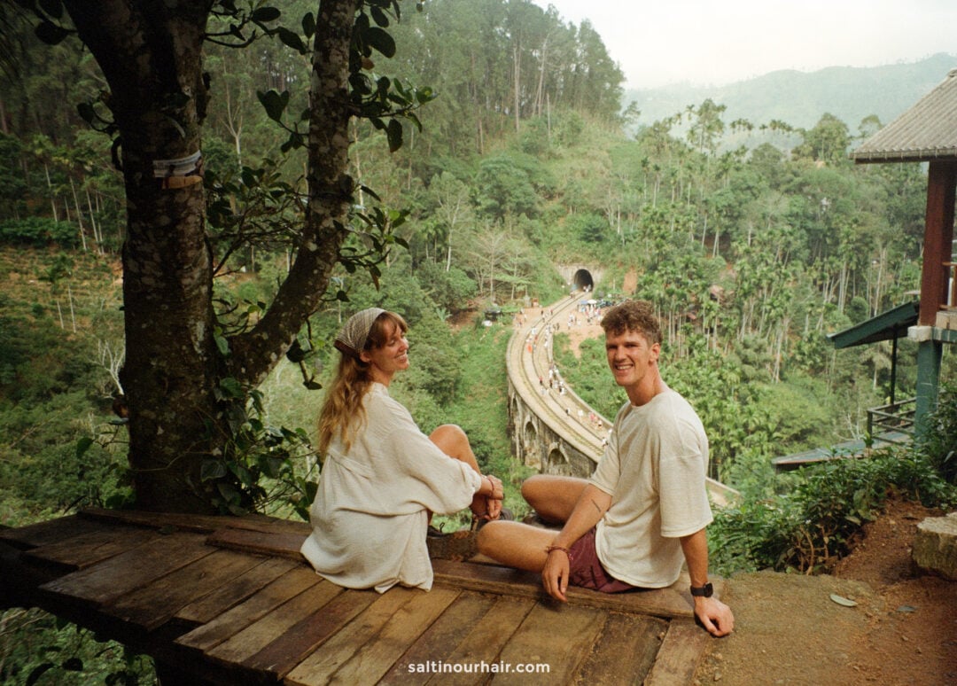 Two people sit on a wooden platform overlooking the curved railway bridge called the nine arch bridge of Ella, Sri Lanka, surrounded by lush greenery and hills.