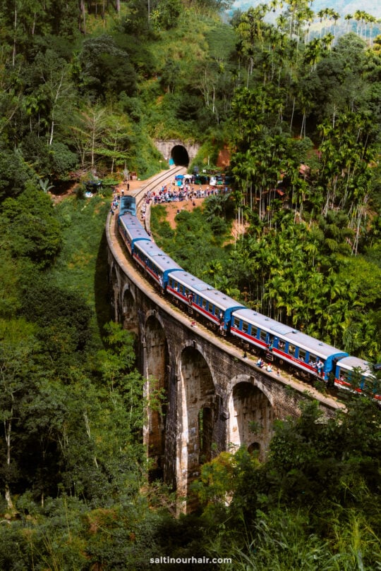 A blue train crosses a curved stone bridge called the nine arch bridge in Ella Sri Lanka, surrounded by lush green forest, with people standing along the tracks and tunnel in the background.