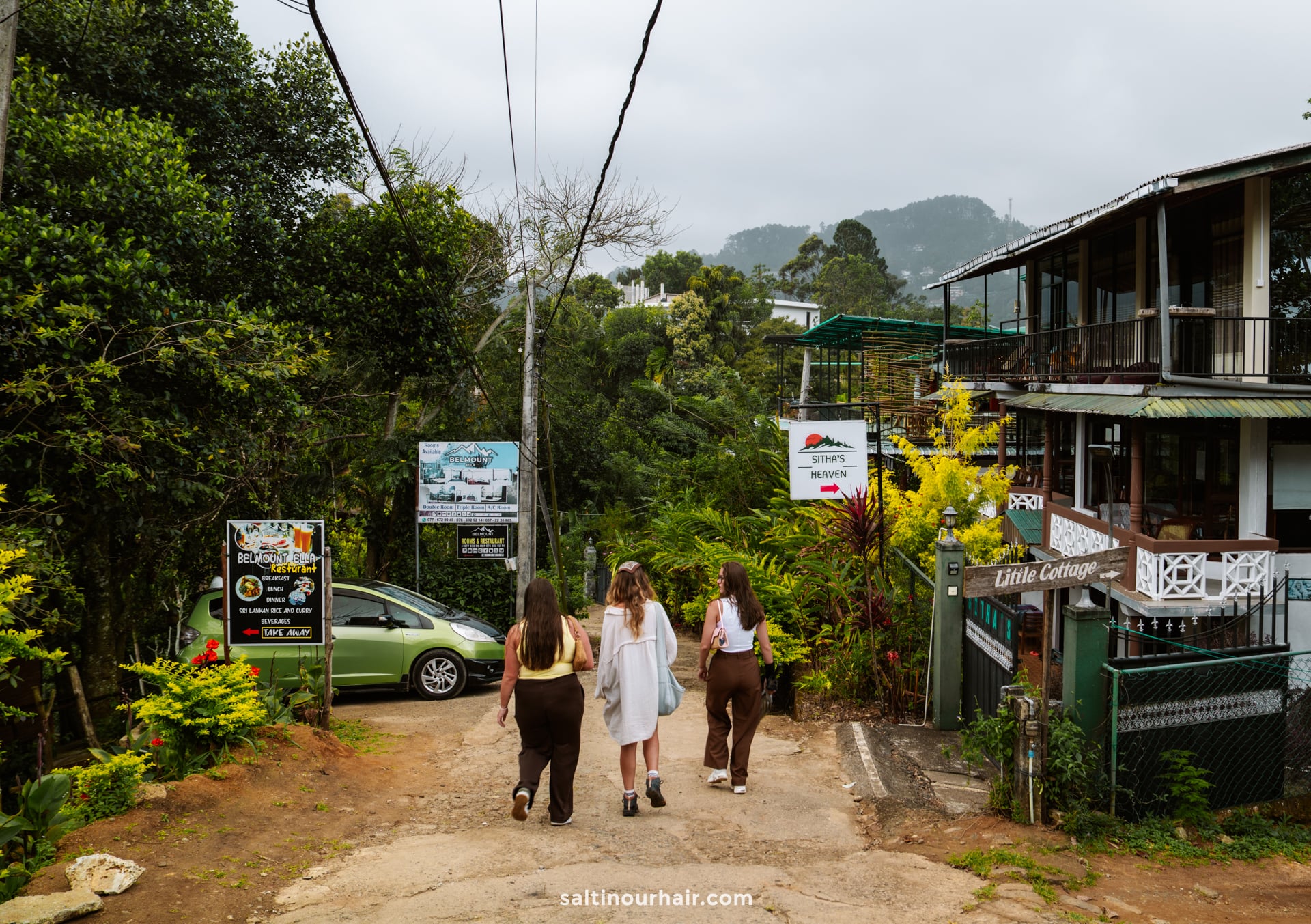 Three women walk down a path toward a green car and several signs outside hillside buildings, surrounded by lush greenery and mountains in the beautiful region of Ella, Sri Lanka.