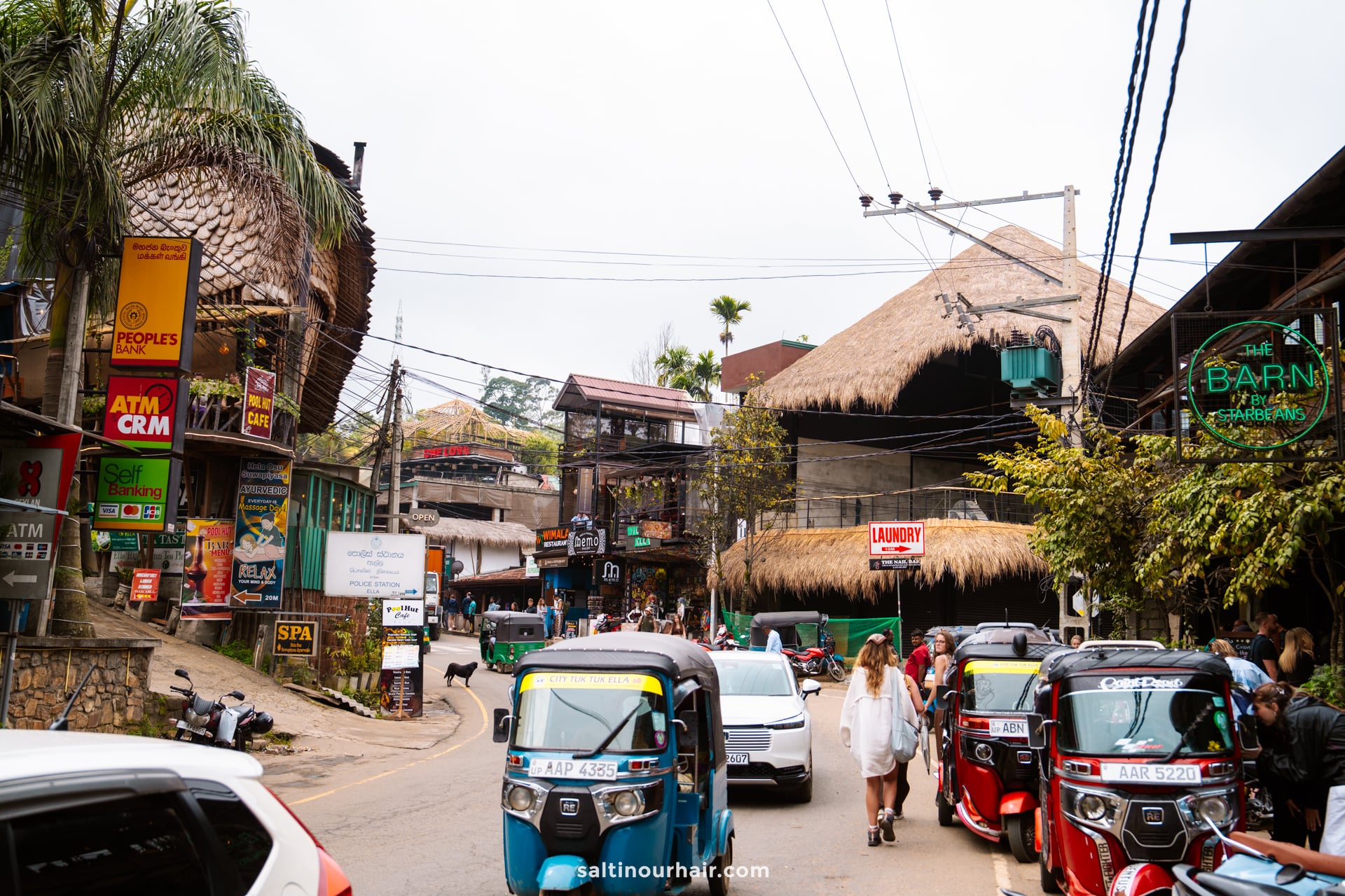 Busy street scene in Ella Sri Lanka, with tuk-tuks, pedestrians, shops, restaurants, and buildings with thatched roofs. Signs and power lines crisscross overhead in this lively tropical town.