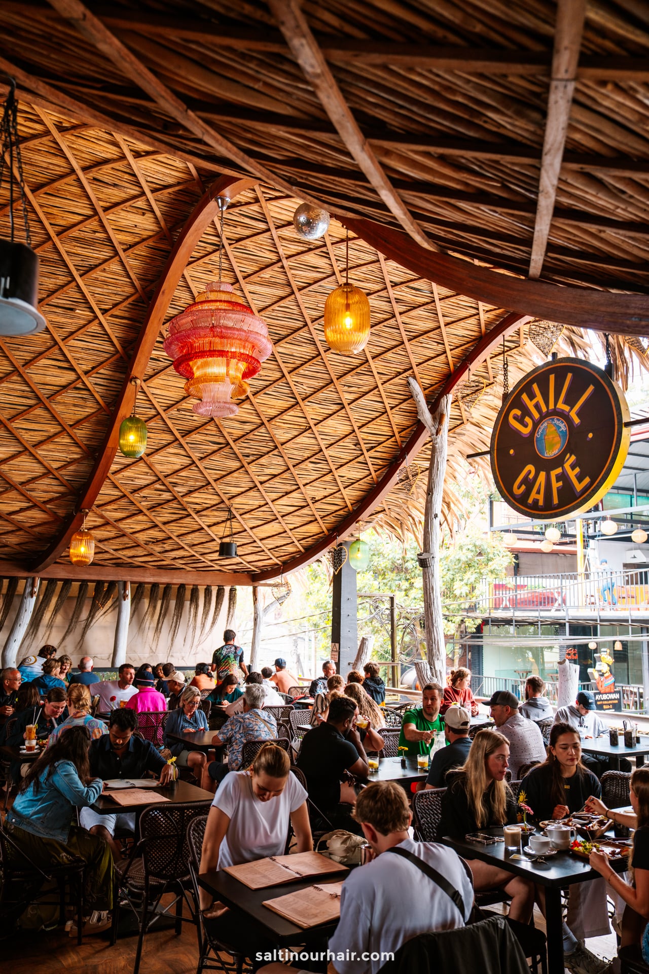 A busy caf&eacute; in Ella, Sri Lanka, with people dining under a large thatched roof, colorful hanging lamps, and a Chill Caf&eacute; sign overhead.