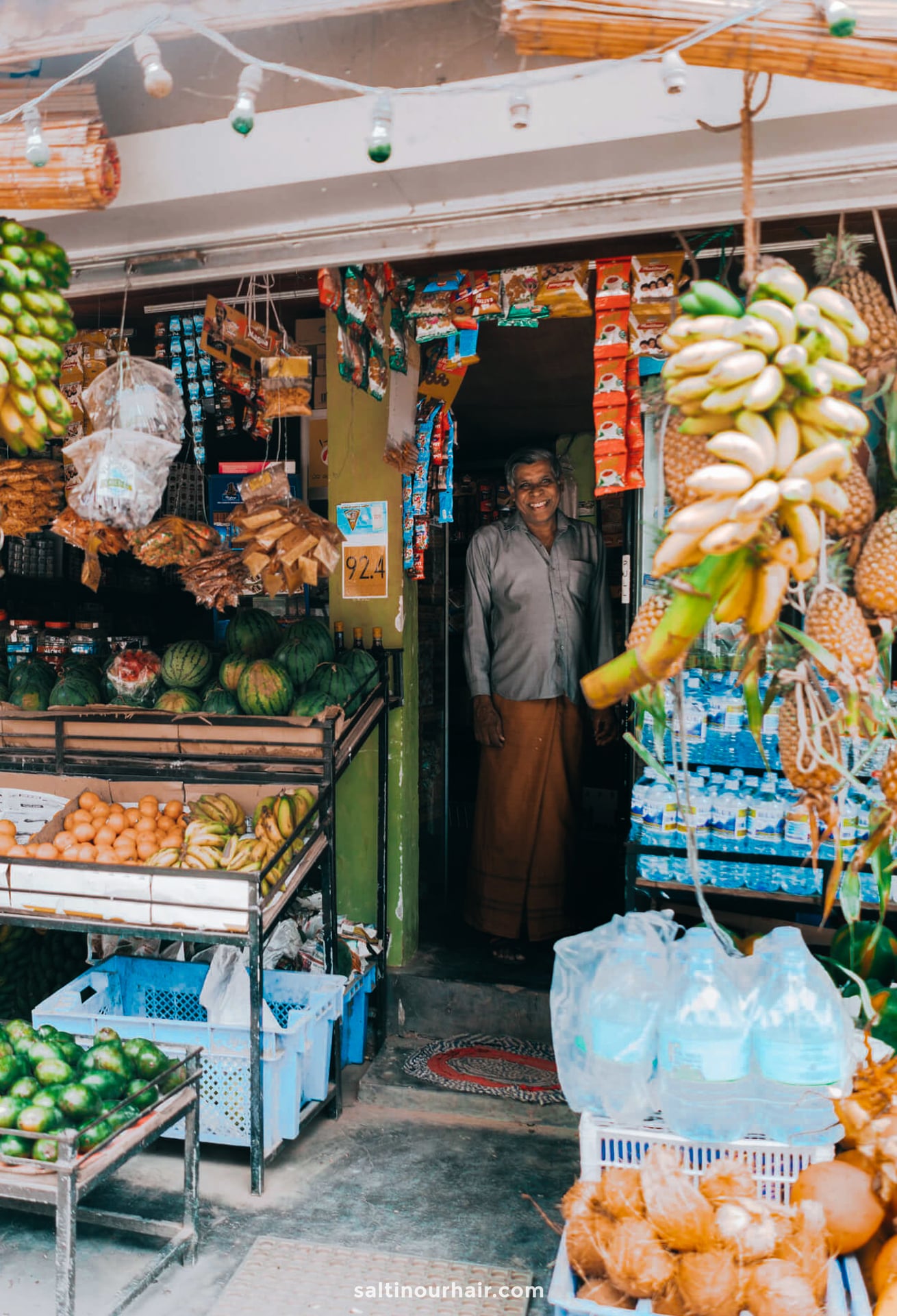 A man stands smiling in the doorway of a small Ella Sri Lanka produce and grocery shop with fruits, vegetables, and packaged goods displayed outside.