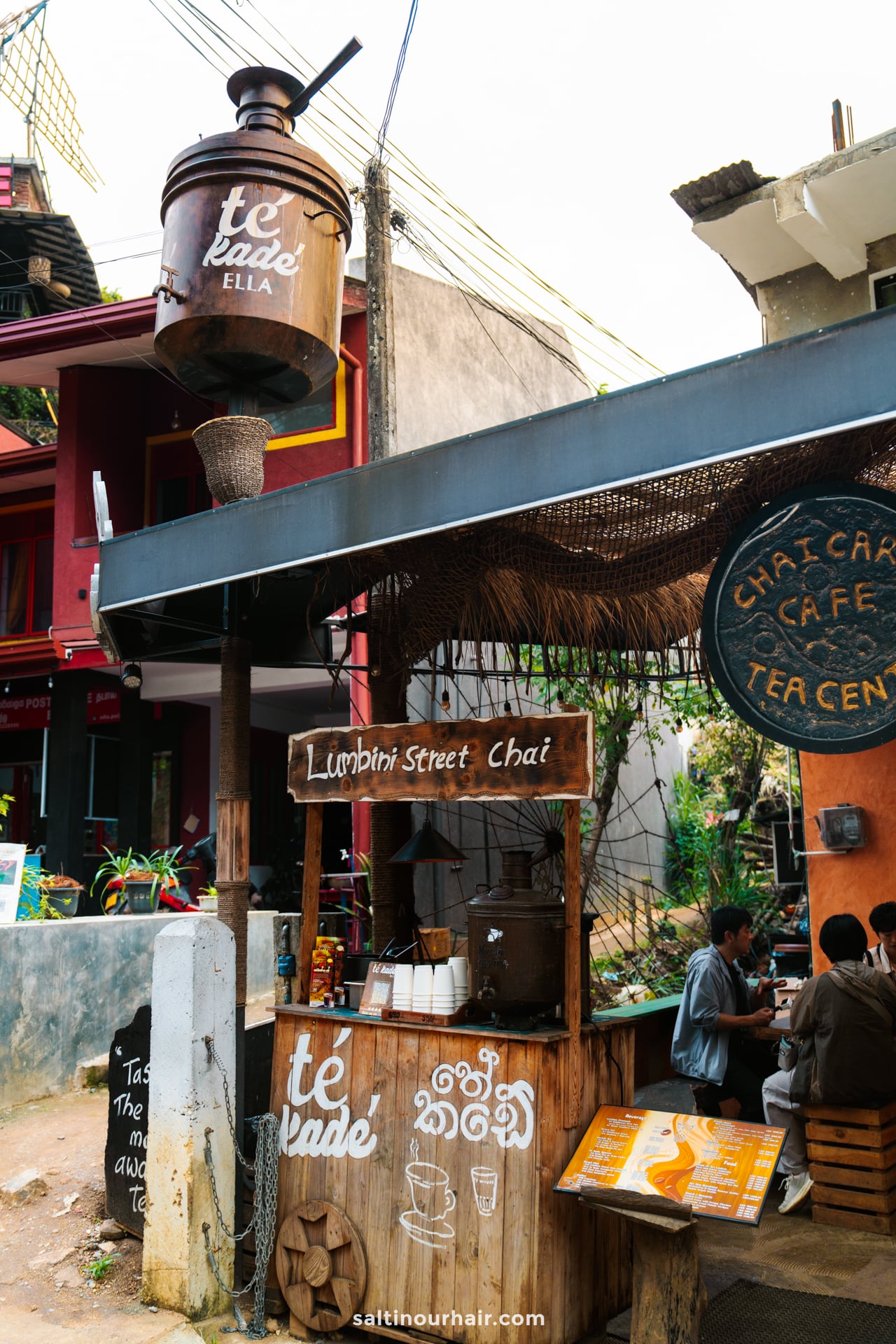 Outdoor chai tea stand with a large kettle sign, wooden counter, and people ordering drinks; set against the vibrant buildings and colorful signage of Ella, Sri Lanka.