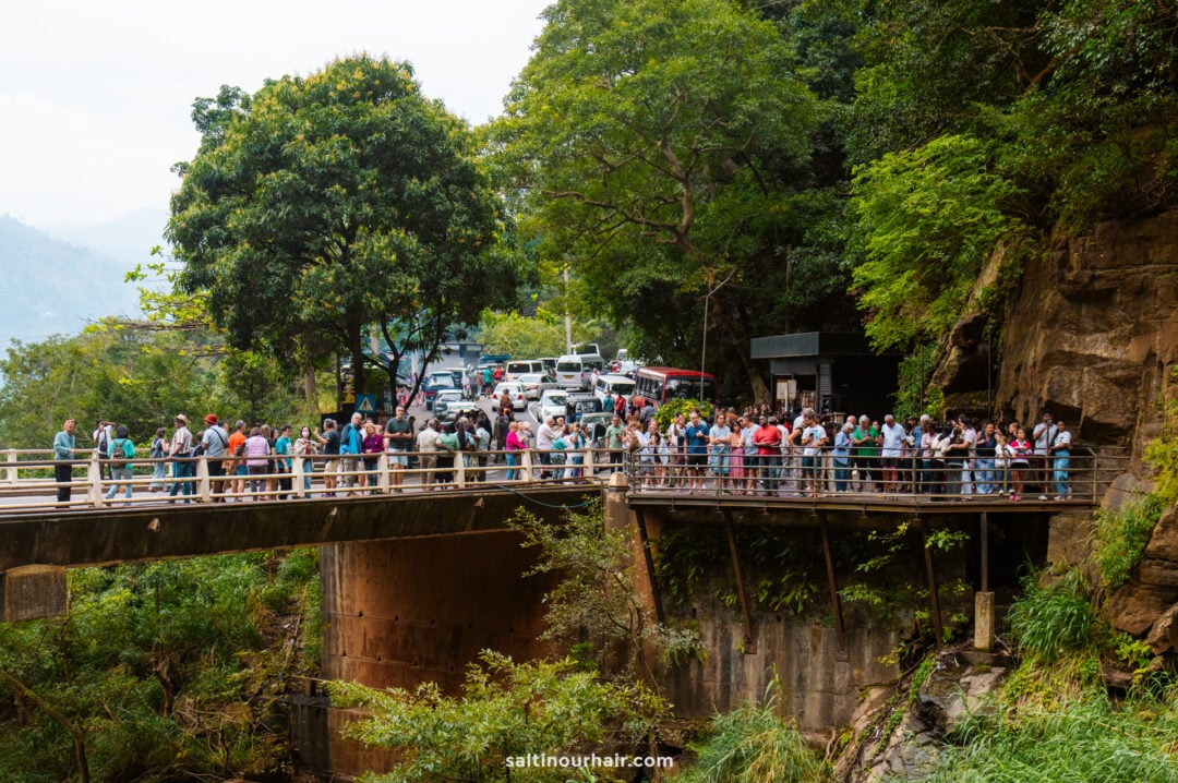 A large group of people stand on a bridge at the Ravana Falls viewpoint, surrounded by trees and parked vehicles, waiting to enter a natural area.