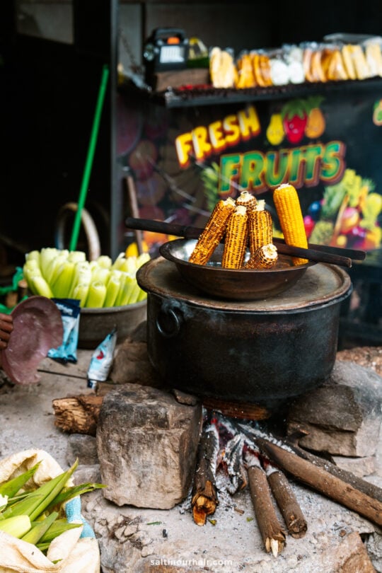 Grilled corn on the cob being cooked over an open fire on a metal pot in Ella, Sri Lanka, with fresh corn and a Fresh Fruits sign in the background.