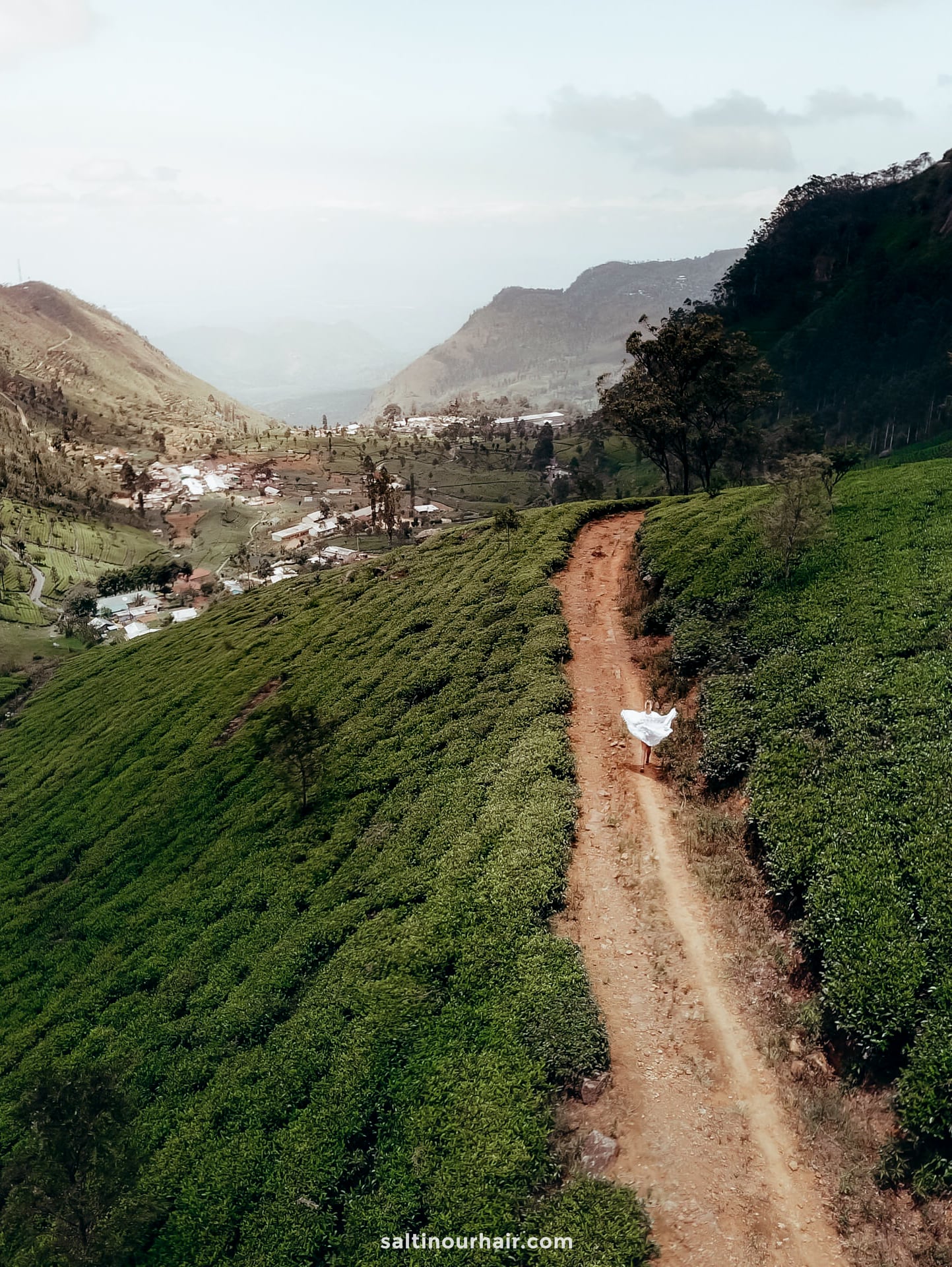 A dirt path winds through the tea plantations of haputale near Ella, Sri Lanka, toward a distant village, with a person in white walking along the path under a partly cloudy sky.