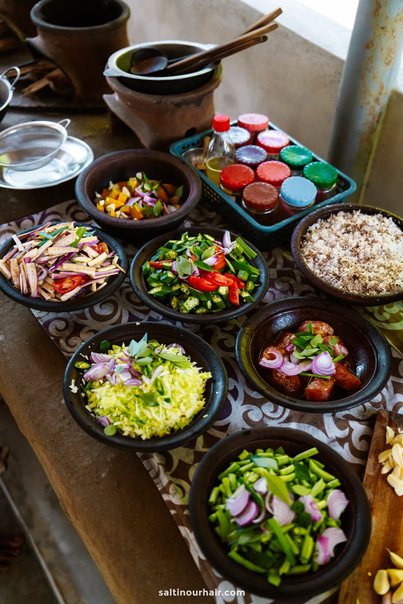 Various bowls filled with chopped vegetables, herbs, and rice are arranged on a patterned surface&mdash;capturing the vibrant flavors found in a cooking class in Ella, Sri Lanka, with spices and condiments in the background.