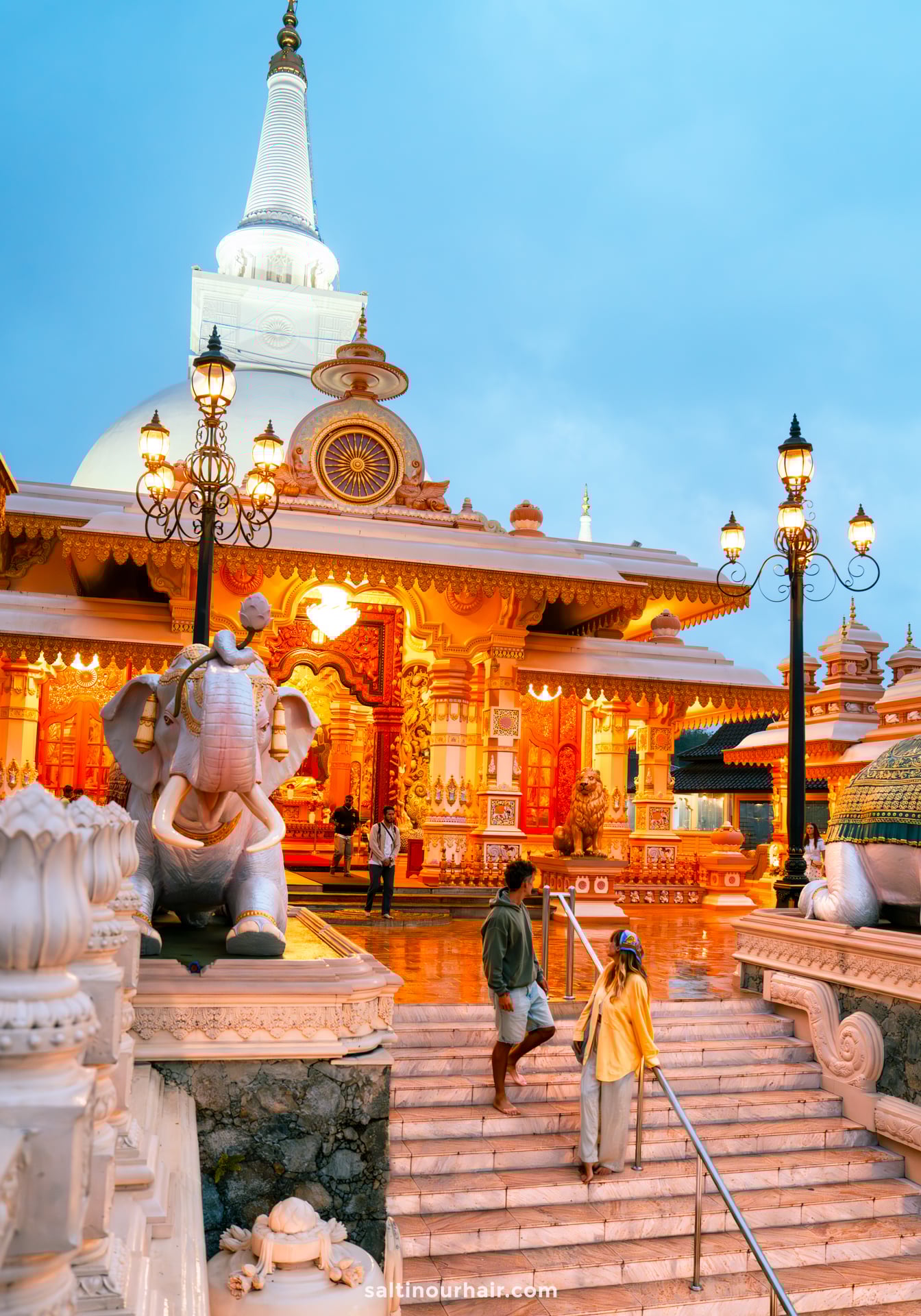 Two people stand near the steps of Mahamevnawa Buddhist Monastery in Ella, Sri Lanka, surrounded by statues and ornate architecture, under a cloudy evening sky.