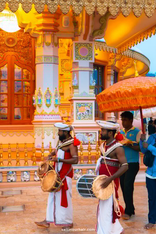 Two traditionally dressed drummers play instruments while walking in a procession near the brightly decorated Mahamevnawa Buddhist Monastery, accompanied by people holding orange umbrellas.