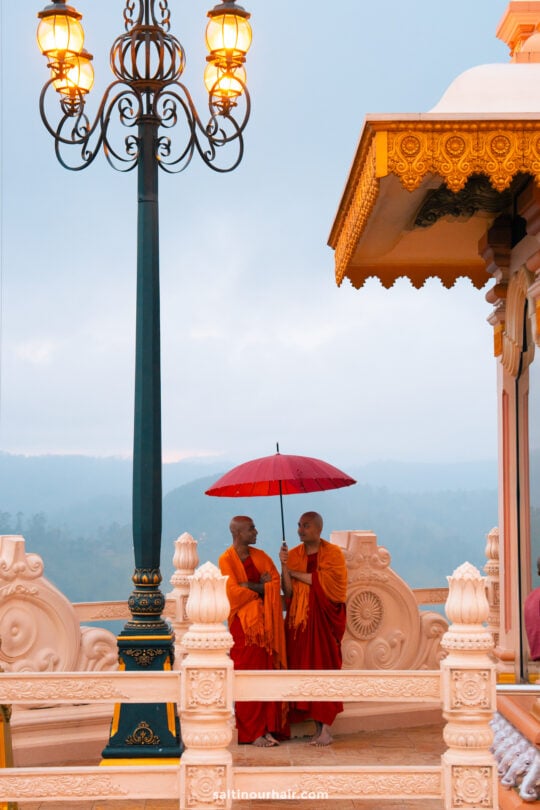 Two Buddhist monks in orange robes stand under a red umbrella near an ornate temple structure and decorative lamp post at Mahamevnawa Buddhist Monastery, with majestic mountains visible in the background.