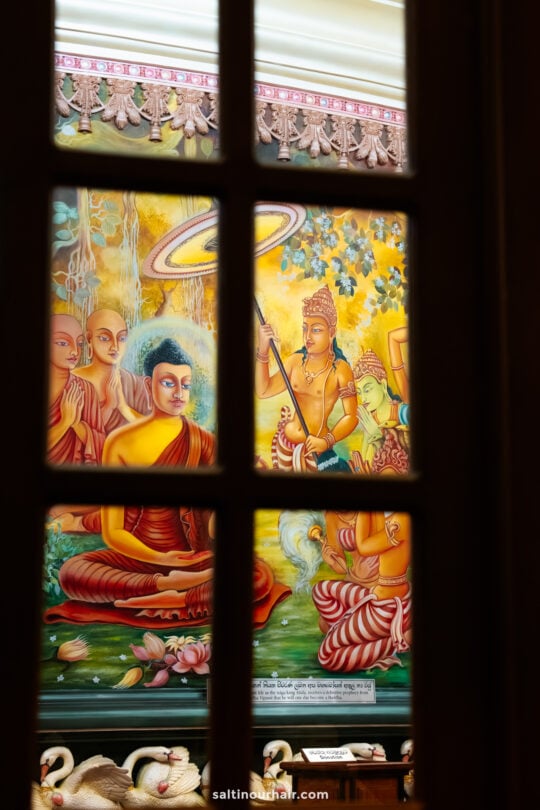 A colorful mural depicting Buddha and attendants is seen through a window with wooden frames in Ella, Sri Lanka. Swans and lotus flowers are visible at the bottom of the serene scene.