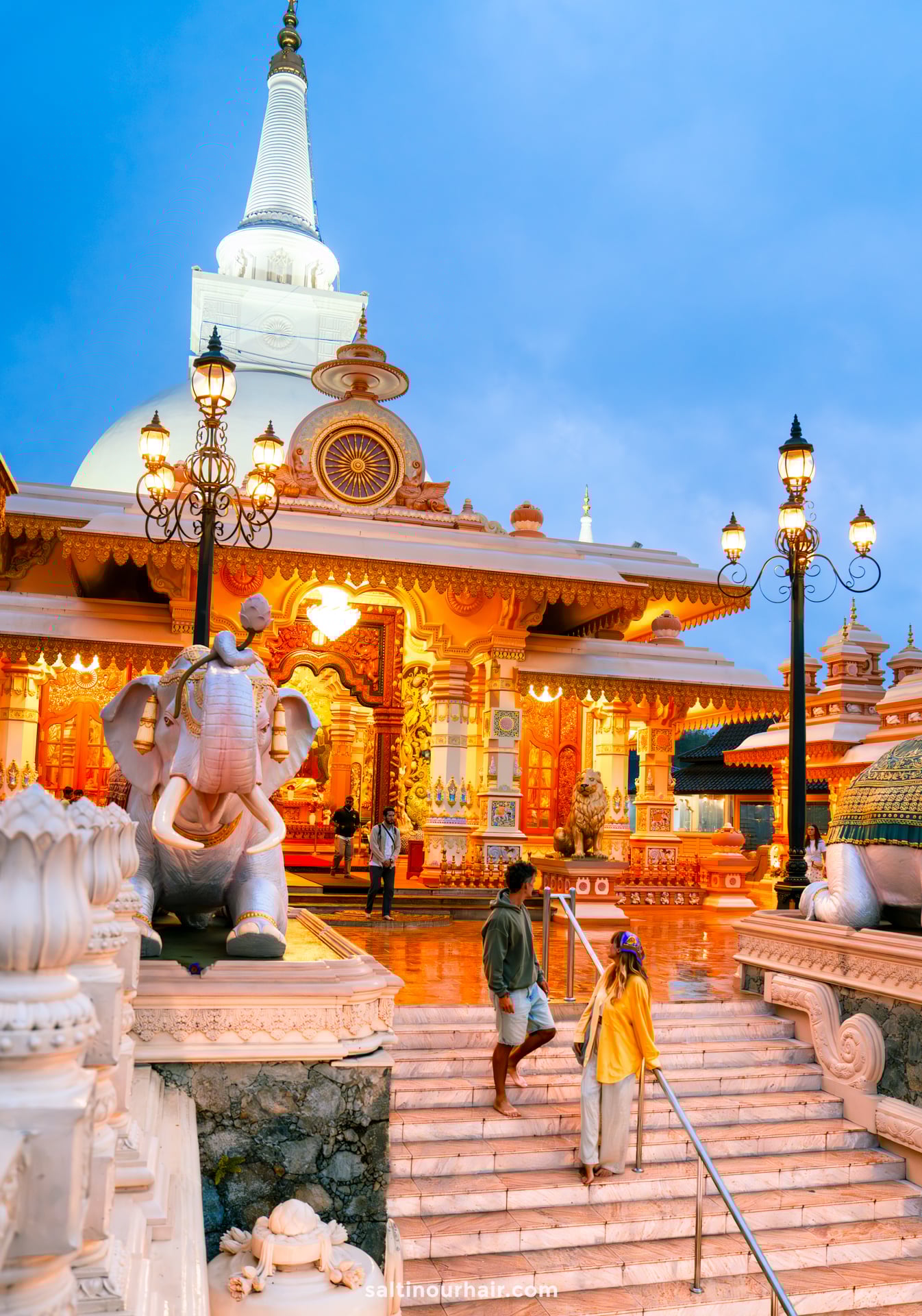 Two people stand at the steps of a brightly lit temple in Ella, Sri Lanka, featuring statues of elephants and ornate decorations, with a large white stupa in the background at dusk.