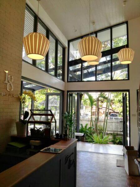 Modern lobby at 18 In Town Homestay Chiang Mai features large windows, pendant lights, potted plants, and a reception desk, opening to a view of the garden and parked car—perfect for your Chiang Mai accommodation.