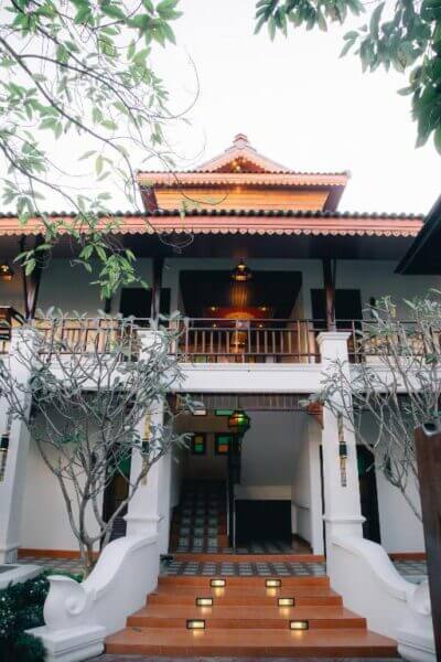 Two-story i Lanna House with a traditional decorative roof, white columns, a central staircase with lights, and surrounding trees—bringing the charm of Chiang Mai to life.
