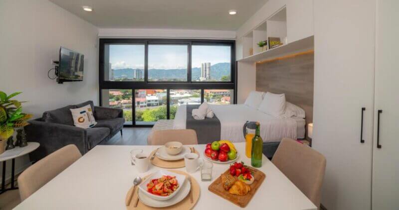 Modern studio apartment in San Jose with a double bed, small sofa, dining table set for breakfast, large window offering city views, and minimalist decor—perfect for a cozy stay at Train Suites.