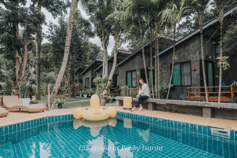 A woman sits on a bench with a suitcase near a swimming pool with a yellow duck float at Family House Pai, a welcoming family guesthouse surrounded by trees and charming bungalow-style buildings.