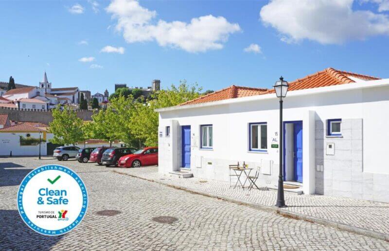 A row of white houses with blue doors sits on a cobblestone street in Óbidos, with parked cars, trees, and a lamppost under a partly cloudy sky. A Clean & Safe badge highlights Mood Lodging’s commitment to guest safety.