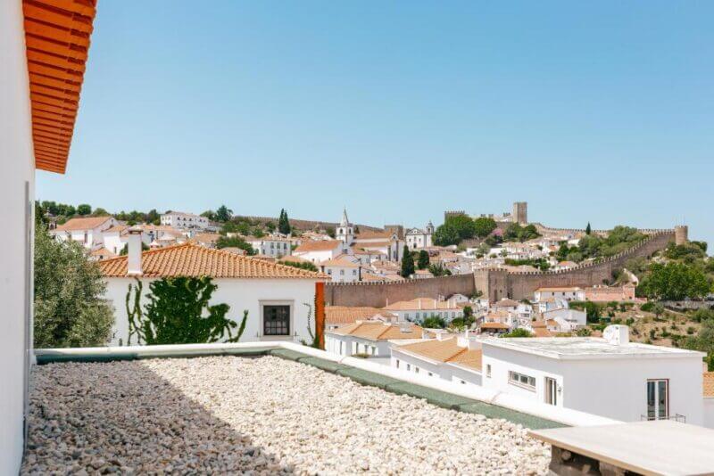 View of Obidos, featuring white buildings, red-tiled roofs, a stone wall, and the iconic Retiro do Castelo castle tower under a sunny sky.