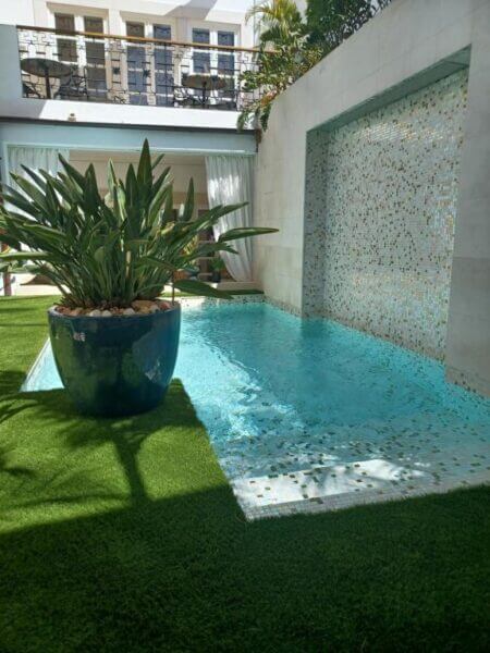 Small pool with tiled waterfall feature at Villa Zawaia B&B Lagos, a potted plant on artificial grass, and a building with balconies in the background.