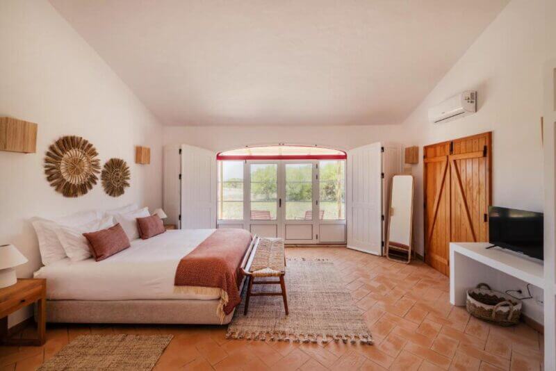 Bright bedroom at Monte da Bemposta with a large bed, neutral decor, woven wall art, a wooden sliding door, a TV on a white console, and double doors leading to an inviting outdoor area.