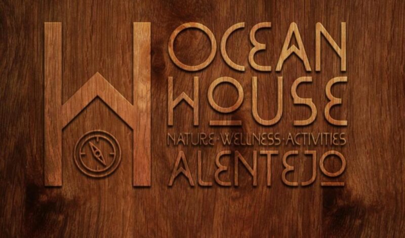 Wooden sign with engraved text: Ocean House Porto Covo Alentejo and Nature - Wellness - Activities, featuring a compass symbol on the left.
