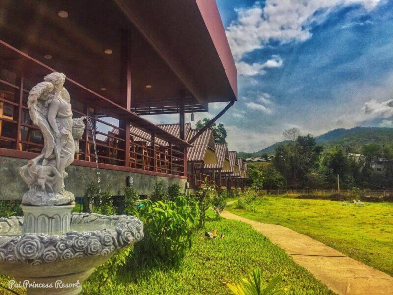A stone angel fountain stands near a garden path beside the Pai Princess Resort’s wooden building, with mountains and a partly cloudy sky in the background.