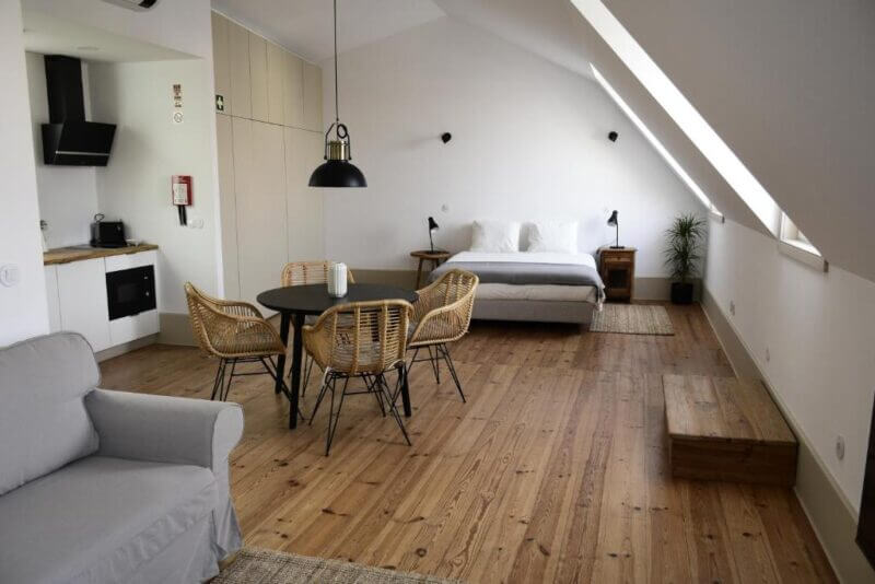 A modern Venezapartments studio in Aveiro featuring a bed, round dining table with four chairs, kitchenette, sofa, and wooden flooring under a sloped ceiling with natural light.