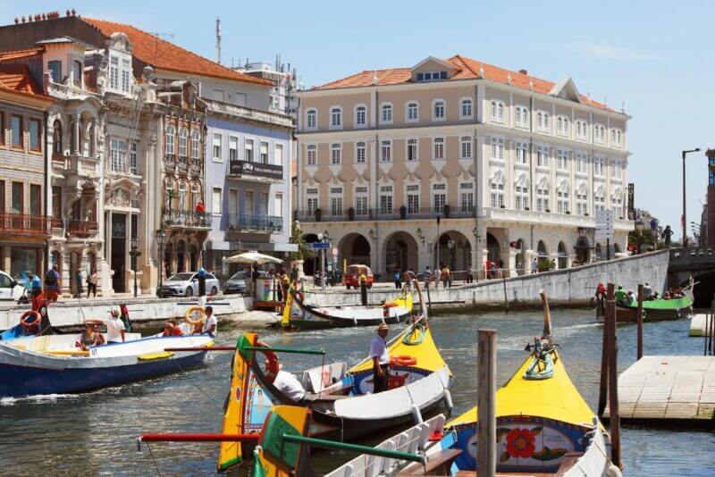 Colorful boats float on a canal lined with ornate historic buildings, including the elegant Hotel Aveiro Palace, on a sunny day, with people visible on the boats and along the water’s edge.