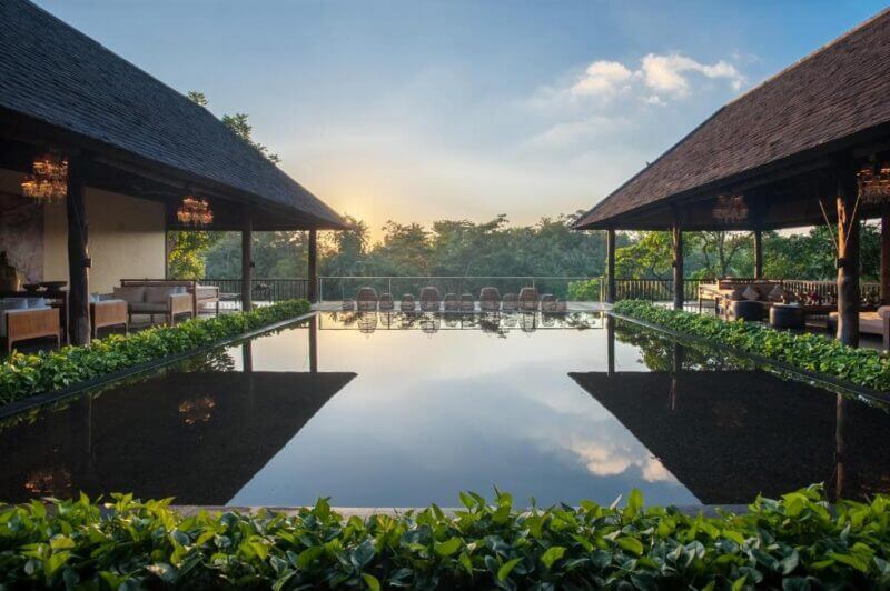 A rectangular reflecting pool at Komaneka Ubud is surrounded by greenery and open-air wooden pavilions with seating, set against a backdrop of trees at sunset.