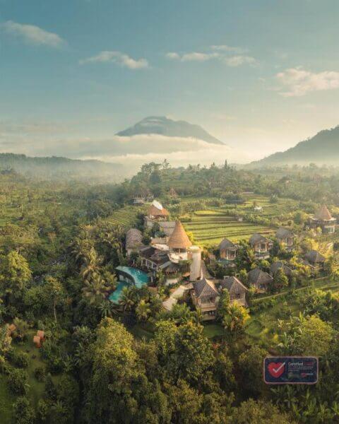 Aerial view of a lush green landscape in Sidemen Bali, with villas, a swimming pool, terraced fields, and a distant mountain under a partly cloudy sky—perfect for a tranquil escape at Wapa di Ume Sidemen Bali resort.