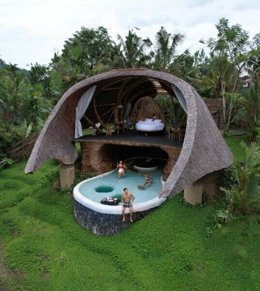A tropical open-air villa at Veluvana in Sidemen features a thatched roof, a loft bedroom, a lower lounge area, and a curved swimming pool where people are relaxing, all surrounded by lush greenery.
