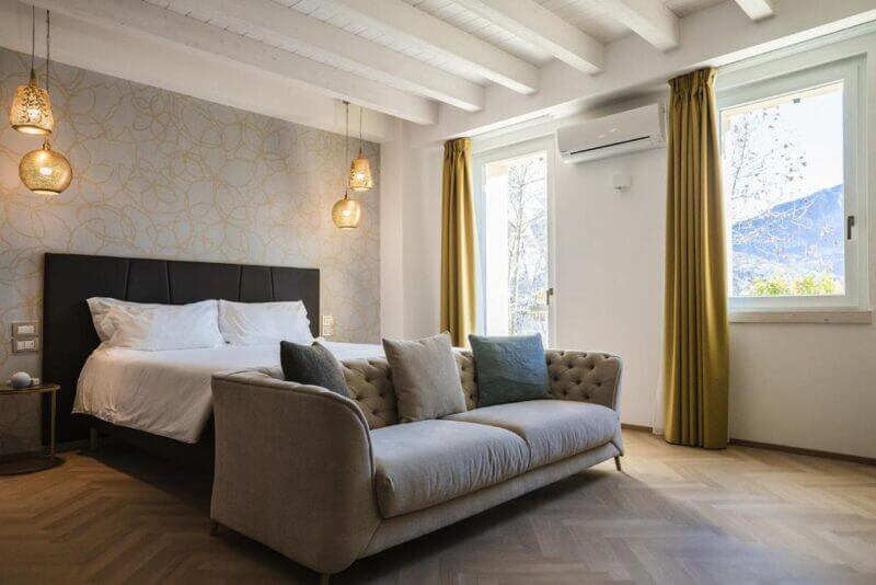 Modern bedroom with a double bed, beige sofa, pendant lights, wood flooring, large windows with yellow curtains, and a mountain view—perfect for those seeking the elegance found in Lake Como Luxury Suites.