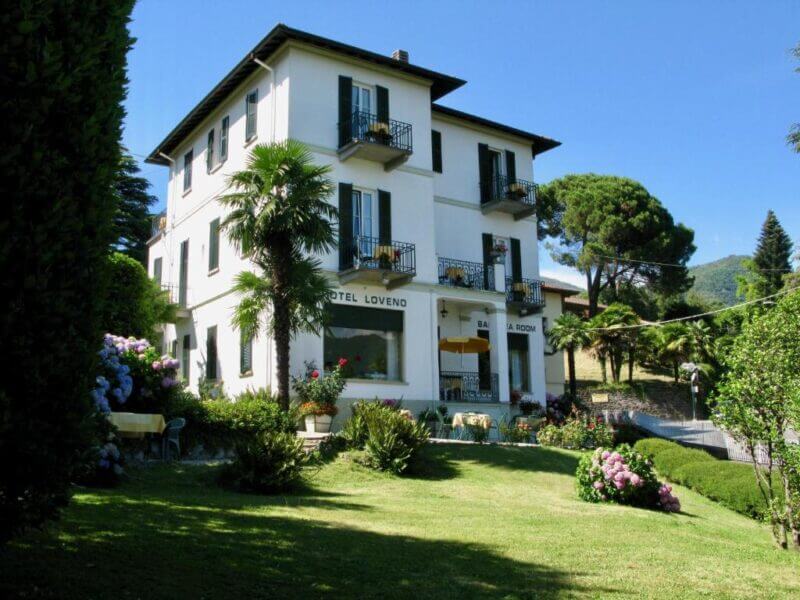 Three-story white Hotel Loveno with balconies, nestled on a green lawn amid trees and shrubs in Menaggio, features outdoor seating and overlooks scenic hills beneath a blue sky.