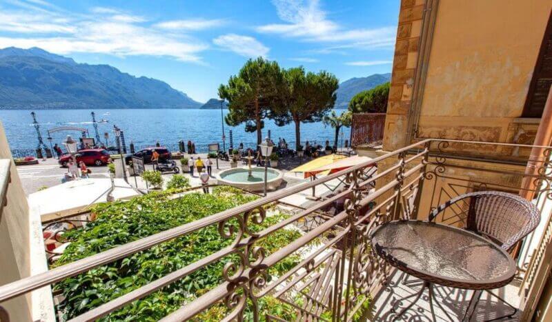 View from a balcony at Hotel Garni Menaggio, overlooking a street, a small fountain, trees, and a lake with mountains in the background under a partly cloudy sky.