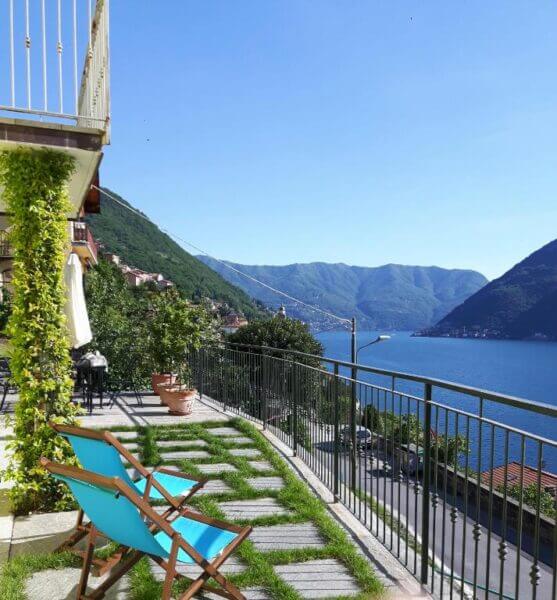 Two blue deck chairs on a grassy terrace in Nesso overlook Lake Como and mountains under a clear blue sky. A metal railing lines the edge, with a house and potted plants nearby, capturing the charm of Balcone dei Limoni.