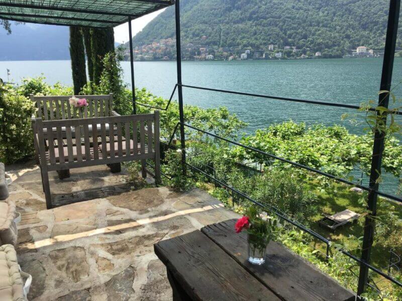 A stone patio at Casa Vacanza Nesso with wooden benches and a table overlooks a lake framed by green hills, while vibrant flowers in vases adorn the table and bench.