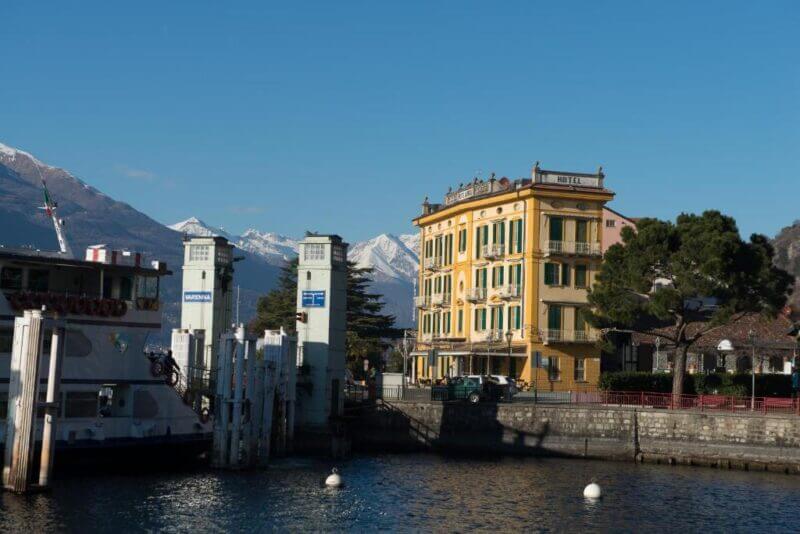 The yellow Hotel Olivedo stands by a waterfront marina in Varenna, with snowy mountains in the background under a clear blue sky.