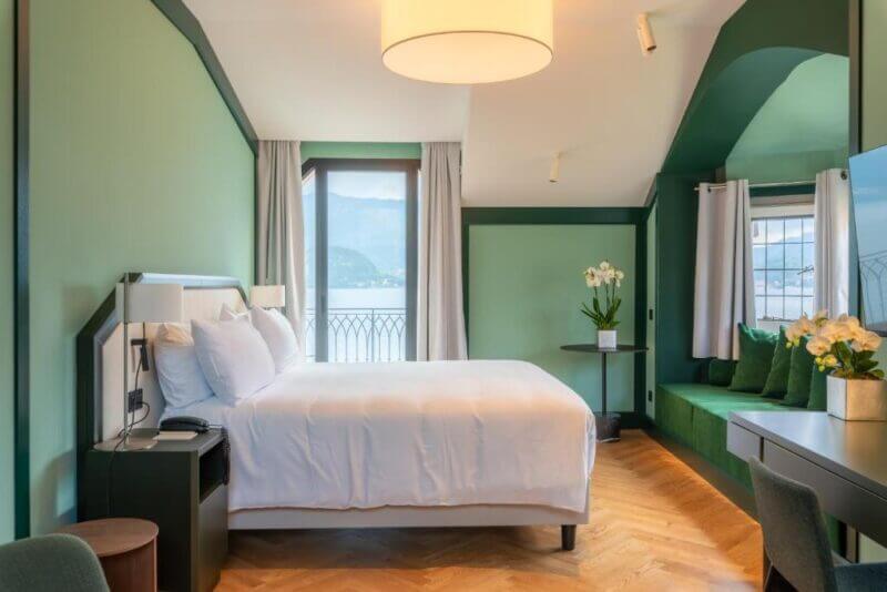Modern hotel room with a large bed, green walls, wood flooring, a window seat, and windows overlooking scenic Lake Como and mountains—experience comfort and stunning views at Hotel du Lac Varenna.
