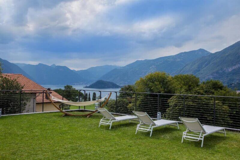 Three lounge chairs and a hammock are set on a grassy lawn at Villa La Rosa Bellagio, offering views of a lake, tree-covered hills, and distant mountains under a cloudy sky.