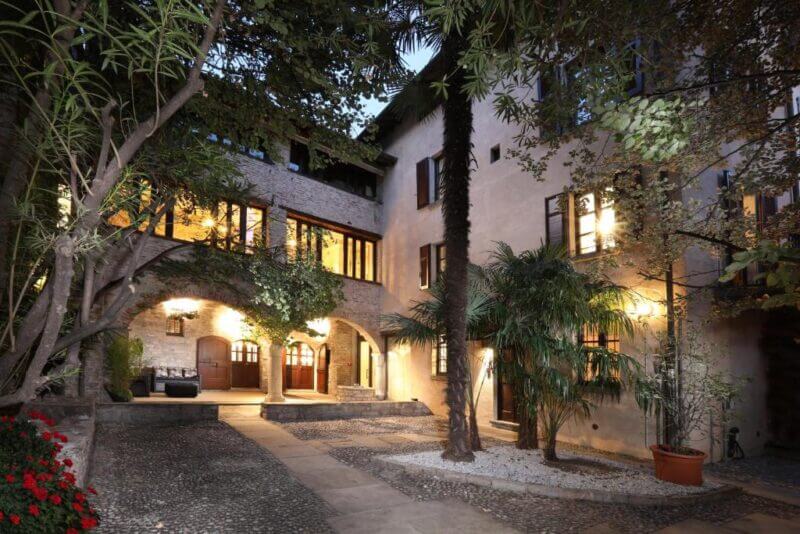 A stone courtyard of Atelier Como with arched doorways, illuminated windows, potted plants, and mature trees at dusk hints at the charm awaiting in its elegant suites.