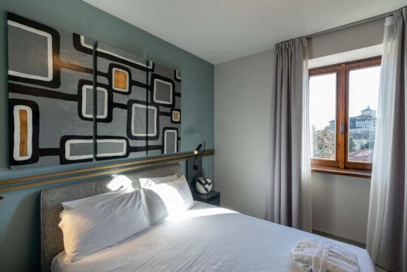 Modern hotel room at Le Funi Hotel with large abstract artwork above a neatly made bed, soft natural light from a window, and a scenic view of Bergamo’s castle outside.