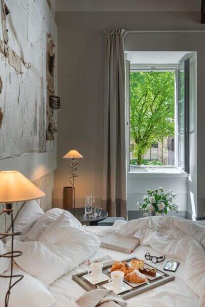 A cozy bed with white linens, breakfast tray, glasses, and books near an open window at GombitHotel in Bergamo, overlooking a green tree and outdoor scene in daylight.