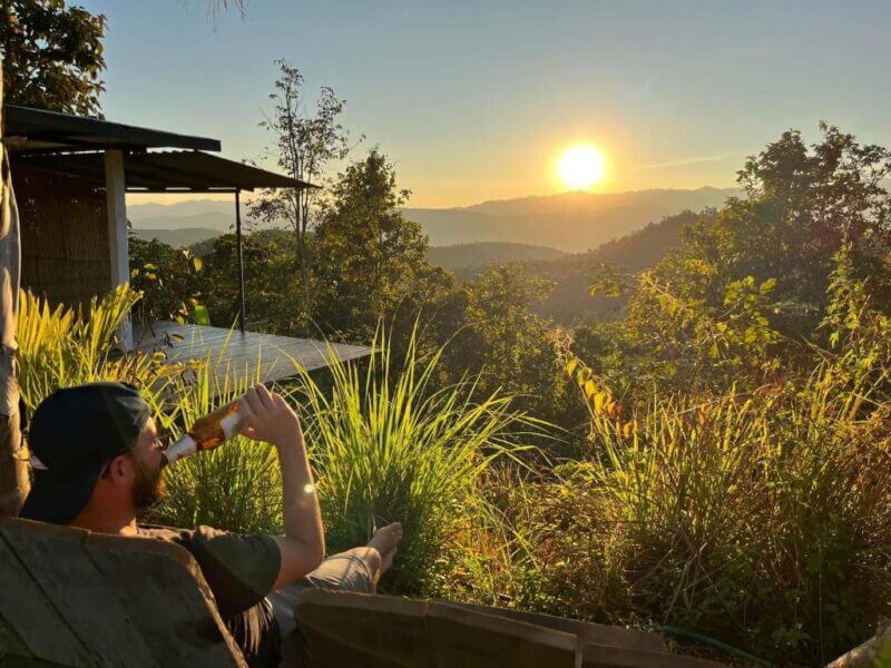 A person sits outdoors in a chair at Monsonkeang, drinking from a bottle, surrounded by greenery, watching the sun set over distant Doi Inthanon mountains.