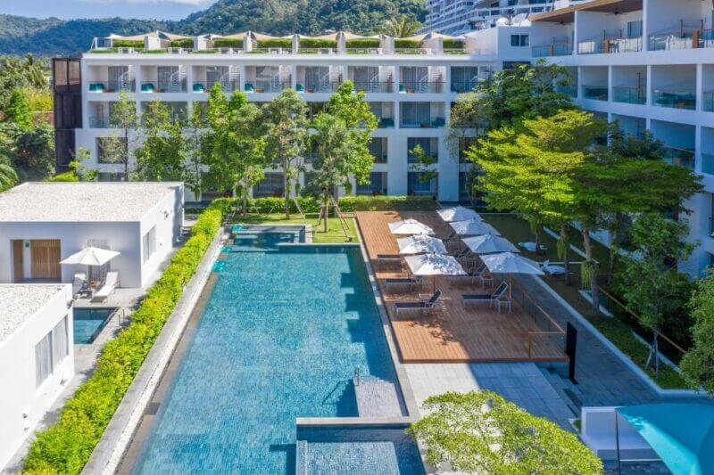 Modern hotel Nap Patong Phuket features balconies overlooking a rectangular outdoor swimming pool, surrounded by trees, lounge chairs, and umbrellas, with scenic hills visible in the background.