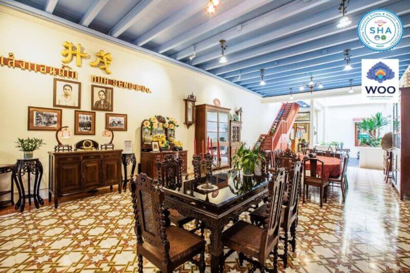 Ornate dining room at WOO Hotel Phuket with carved wooden furniture, patterned tile floor, framed photos on the wall, and plants; blue ceiling beams and a staircase visible in the background.