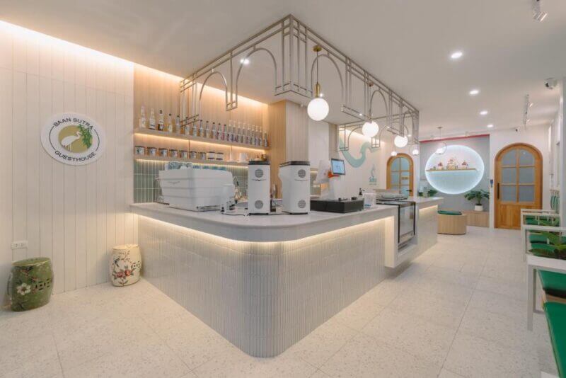 Modern café interior at Baan Sutra Phuket with a light color palette, coffee machines on the counter, shelves with bottles, round pendant lights, and seating along the walls.