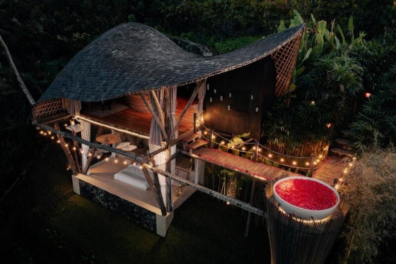 A bamboo villa with a curved roof, open-air living area, string lights, and a round outdoor tub filled with red flower petals, surrounded by dense greenery.