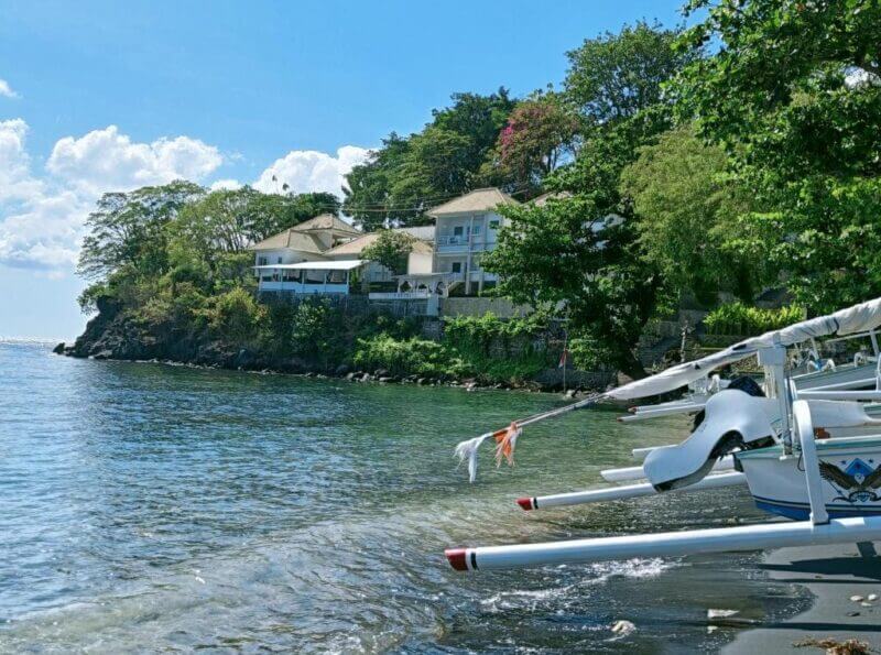 White buildings sit on a tree-covered shoreline above clear water, with traditional boats docked along the beach under a bright blue sky—an inviting scene typical of Bali accommodation like Aquaterrace Amed or a charming Amed hotel.
