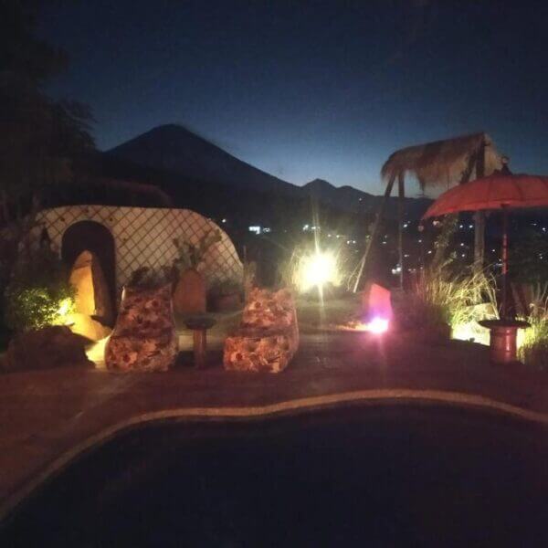 Outdoor seating area by a pool at night in Blue Earth Village, Amed, Bali, featuring two chairs, small tables, decorative lights, and scenic mountains in the background.